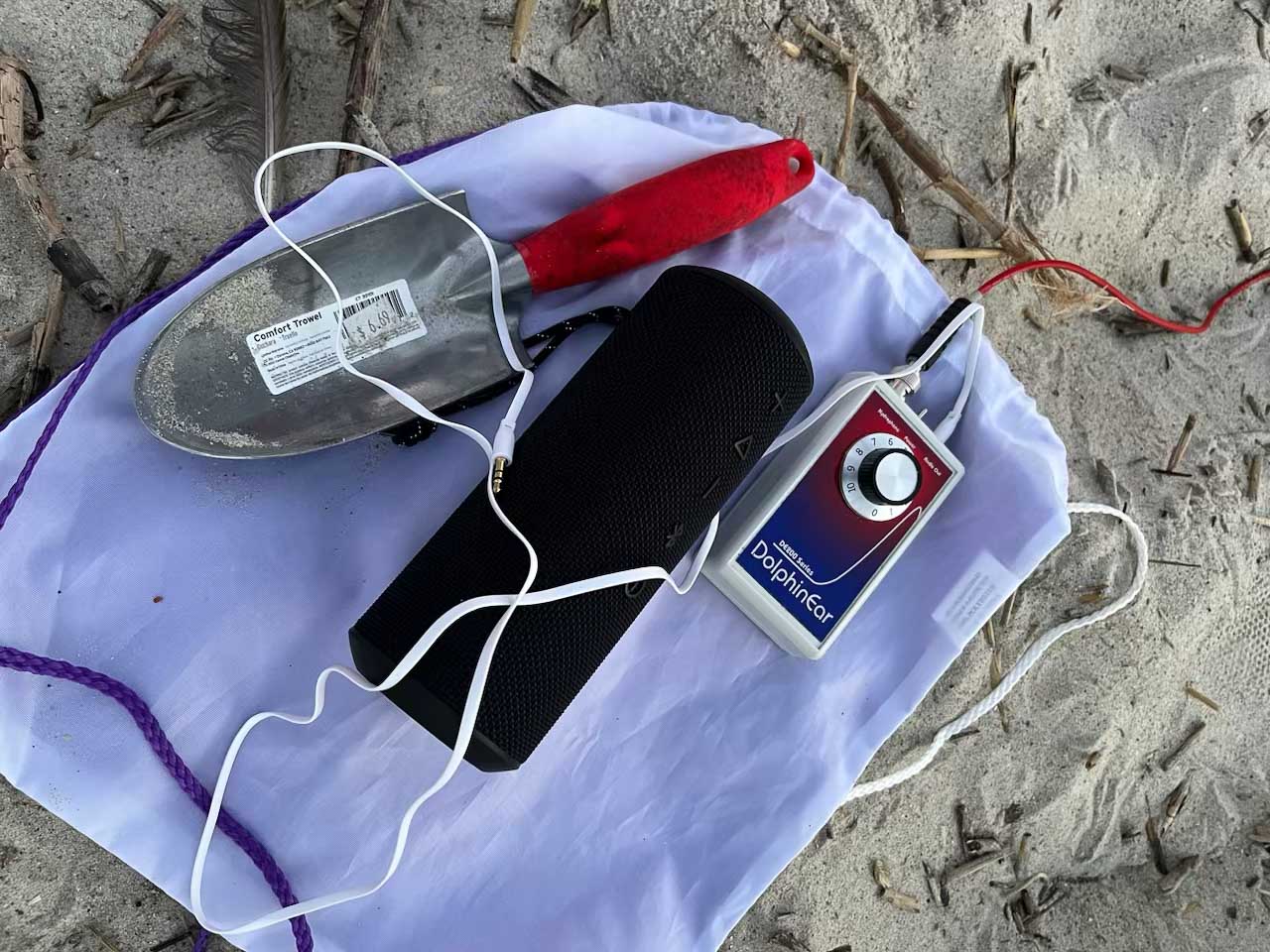 A DolphinEar hydrophone with a bluetooth speaker and a hand trowel sitting on a backpack on the sand.